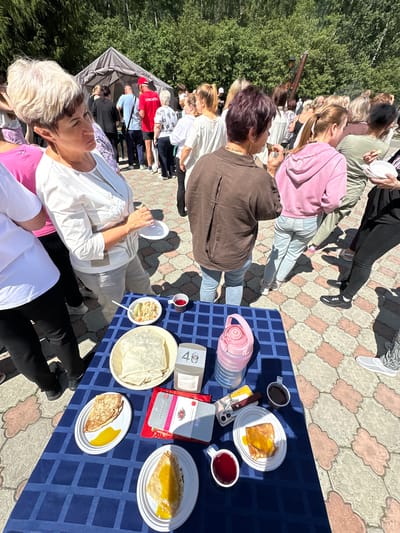 A group of people are gathered outdoors in what appears to be a public event in Duvankul', Russia. The scene is dominated by a large tent in the background, suggesting an organized gathering or festival. In the foreground, a table is set with various food items and drinks, including plates of crepes, cups of red liquid, and a large pink water bottle. Several individuals are visible, some looking towards the food table, while others are engaged in conversation or observing the surroundings. The ground is paved with interlocking tiles, and lush green trees form a backdrop to the event. The overall atmosphere seems casual and social, indicative of a community gathering or celebration.