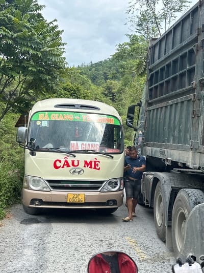 A partially obstructed, medium-shot scene on a road in Bạch Đích Commune, Vietnam, depicts a beige Hyundai passenger van with the inscription "HÀ GIANG - YÊN MINH" and "CẦU MÈ" in red letters on its front windshield, partially obstructed by a large green truck. A man in a blue shirt and grey shorts, wearing sandals, leans towards the front of the van, appearing to inspect the area under the truck's wheels. The truck's tires are visible, indicating its proximity to the van. The van's license plate is visible, reading "23F 000.86". The road surface is asphalt. Green foliage and trees are visible in the background, suggesting a rural or mountainous location. A partial view of a red motorcycle mirror is visible in the right side of the frame.