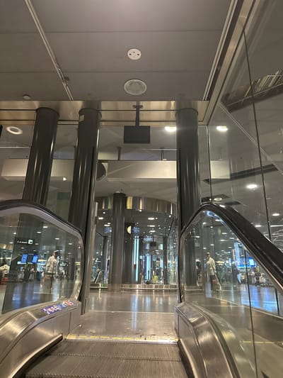 This image shows an interior view of Zurich Airport in Kloten, Switzerland, featuring parallel escalators. The escalators are encased in glass and metal, with black handrails. Reflections of people, signs, and screens are visible in the glass. Gray metal supports are visible. The ceiling is made up of a combination of glass panels. The lighting consists of recessed lights. The floor is grey.