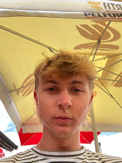 A young, light-haired male, appearing to be in his late teens or early twenties, is depicted in a front-facing, close-up portrait, likely a selfie, taken outdoors in Châtel, France. He has unkempt, light brown or blonde hair and greenish-brown eyes, with a neutral expression and minor facial blemishes on his chin and cheeks. The subject is wearing a striped t-shirt with white, black, and green/beige horizontal stripes. The scene is set beneath a large, yellow commercial umbrella, which prominently displays the reversed branding "PELFORTH" and a bird logo, indicating a French brewery. Visible structural components of the umbrella include its spokes and central mechanism. To the lower left, a portion of a red umbrella or awning is visible behind the subject, alongside a dark-colored roofline. A clear blue sky with white clouds is discernible in the upper left quadrant, beyond the umbrella's canopy, suggesting daytime conditions.