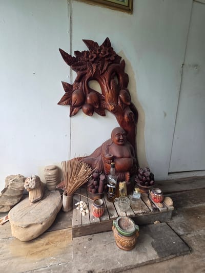In Koh Toch Village, Cambodia, a small shrine is set up on a weathered wooden surface against a white wall. The shrine features a prominent wooden carving of a tree with fruit and flowers, which serves as a backdrop for a seated Buddha statue. In front of the Buddha statue, there's a small collection of items, including a bottle of whiskey, several small ceramic pots with decorative designs, a small glass jar, and a bundle of incense sticks. Scattered around the shrine are various stones, some appearing to be carved or shaped, and a large, flat stone object. The overall impression is of a personal altar or display, likely in an outdoor or semi-outdoor setting given the rustic wooden platform and natural elements.