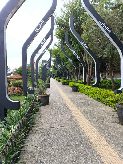 A pathway leads through a park or garden, bordered by black, curved metal structures adorned with Arabic script. Potted plants line the path, which features a tactile paving strip for visually impaired individuals. Lush greenery and trees create a natural backdrop. People are seen in the distance, enjoying the serene environment. The structures cast shadows on the path, enhancing the depth and perspective of the scene.