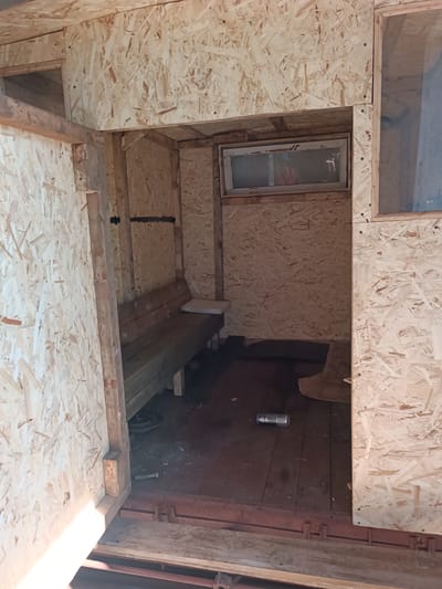 The image shows the interior of a small, unfinished wooden structure, likely a shed or outbuilding, constructed primarily from oriented strand board (OSB). The structure is located outdoors, with glimpses of natural light suggesting it is daytime. The walls, ceiling, and floor are all made of OSB, giving the interior a rough, rustic appearance.
Inside the structure, there is a long, wooden bench built along one wall. On the bench, a white cushion is placed at one end. The floor appears to be made of dark wooden planks. In the middle of the floor, a single aluminum can is visible.
There is a small, rectangular window set into the far wall, with a light-colored frame and dirty glass. The window is positioned high up on the wall. Another opening, possibly a doorway or another window, is visible on the right side of the frame, with a clear plastic sheet serving as a makeshift windowpane.
The entrance to the structure is on the left, and the framing for the doorway is visible, constructed from rough-hewn wooden beams. The overall impression is one of a construction site or a recently built, but not yet completed, functional space. No people are visible in the image. The context of Vuhlehirsk, Ukraine is not evident from the visual details alone.