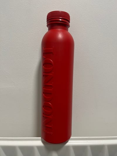 The scene, located in the city of Bath, United Kingdom, displays a vertical, matte red water bottle positioned against a textured off-white wall. The bottle features a matching red screw-top cap with horizontal ridges. The word 'LONDON' is embossed vertically downwards in capital letters on the bottle's front surface. Below this, smaller, faint raised text reading 'bottle up' is partially discernible. The bottle casts a soft shadow to its right on the wall. It rests on a white, horizontally slatted structure, consistent with a radiator, which spans the lower portion of the image. No people or other active subjects are present.
