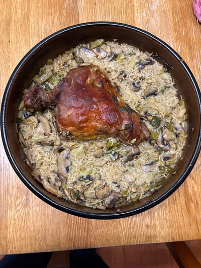 A round, black, ceramic dish filled with rice, mushrooms, and leeks, with a roasted lamb shank on top, sits on a wooden table. The photo appears to have been taken from directly above. The location is likely somewhere in the United Kingdom, possibly Oswestry. No people are visible in the image.