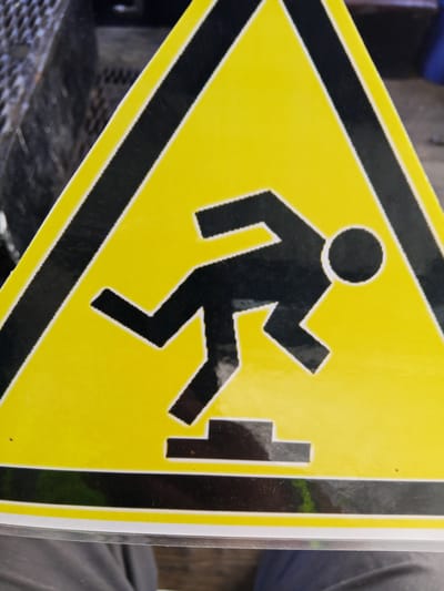 A yellow triangular warning sign with a black border depicts a stylized black figure stumbling on steps. The figure appears to be in mid-fall, with one leg extended and the other bent, and its arms are out to the sides. The sign is mounted on a dark, textured surface, possibly a piece of equipment. The lower portion of the image shows a person's lap covered in gray fabric, suggesting the sign is at waist height or lower. The environment appears to be outdoors, as indicated by the lighting and background elements, though specific details of the Raygorodok, Kazakhstan location are not discernible from the image itself.