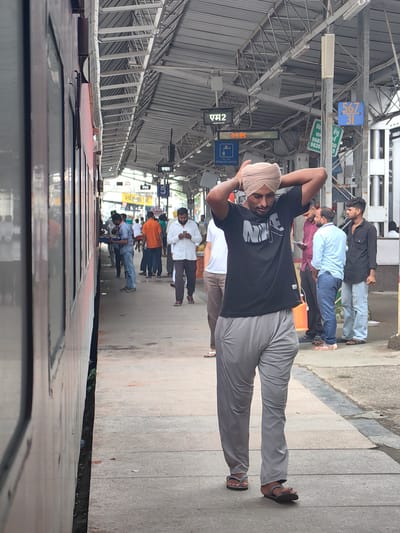 A medium-shot photograph shows a man wearing a turban and a black t-shirt with a Nike logo, walking on a train platform. He is in the foreground, facing away from the camera, with his hands on his head. His pants are grey, and he is wearing flip-flops. In the background, other people are visible on the platform, some standing and some walking. A train is parked on the left side of the image. The environment appears to be a train station in Khandwa, India, with a covered platform and various signs and structures typical of a railway station. The lighting suggests it is daytime.