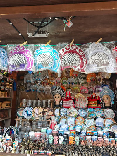A brightly lit market stall in Uglich, Russia, displays a variety of souvenir items. At the top, a row of colorful, ornamental headdresses hangs from clothespins, above a second layer of smaller headdresses. Below this, mannequin heads wearing decorative headdresses are visible. The middle ground features a shelf of decorative plates and glassware, including mugs. The foreground is filled with small figurines and souvenirs.