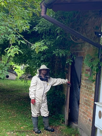 In Albertowsko, Poland, a person wearing a full beekeeper's suit, including a veiled hat, light-colored protective clothing, red gloves, and dark rubber boots, stands outdoors. The individual is positioned near an open wooden door set into a brick building. The person's right hand rests on the edge of the door, and they are looking towards the camera. The environment is lush and green, with dense foliage from trees and bushes surrounding the area. In the background, a small wooden structure with a circular opening, possibly a birdhouse or part of a beekeeping setup, is visible amongst the greenery. A dark downspout runs vertically along the side of the brick structure. The ground is covered in grass and fallen leaves.