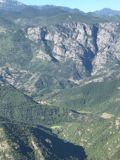 The image is an aerial view of a mountainous, forested landscape in the vicinity of Kućišta, Montenegro. The terrain is characterized by steep, rocky cliffs and rolling hills densely covered with dark green vegetation. A small cluster of buildings, possibly a village or a hamlet, is visible nestled in a valley amidst the trees. Further into the distance, a bright blue body of water, likely a lake or a reservoir, is partially visible. In the upper right section of the image, a series of geometric shapes, possibly agricultural fields or solar panels, are laid out on a hillside. The sky is clear and blue.