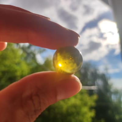 A close-up shot captures two fingers holding a transparent, greenish-yellow marble. The marble is illuminated by bright sunlight, creating a strong glint and lens flare within its center. Tiny specks are visible within the marble. The background is softly blurred, showing lush green foliage and a bright sky with white clouds and the sun. The overall lighting suggests a sunny day, possibly late morning or afternoon. The image appears to be taken outdoors, with the natural light and greenery indicating a possible park or garden setting. There is no visible text in the image.