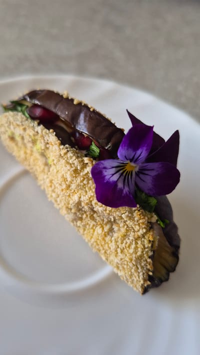 The image displays a close-up, overhead shot of a single, elaborately prepared food item presented on a white plate. The food item appears to be an appetizer or small dish, consisting of what looks like a piece of breaded eggplant or zucchini, possibly stuffed with dark red ingredients, like pomegranate seeds, and garnished with a vibrant purple viola flower and green herbs. The breading is a light golden-brown and finely crumbed. The background is a neutral, textured surface, likely a countertop or table, in a muted gray tone. The lighting is soft, highlighting the textures and colors of the food. The location of Krauja, Latvia is not depicted in this image, as the scene focuses solely on the food presentation.