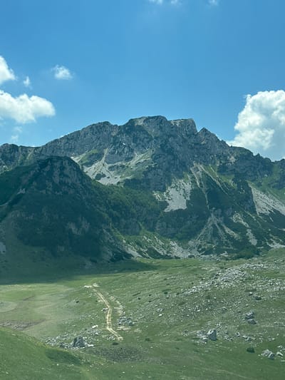 The image shows a steep, rocky mountain slope covered in patches of green vegetation and scattered rocks. The mountain face is rugged, with visible strata and crevices. A winding dirt path is visible on the grassy slope, ascending the mountain. The sky is blue with scattered white clouds. The scene appears to be set in a mountainous region, likely during daylight. The image is oriented vertically, with the mountain dominating the right side of the frame and the sky on the left. The presence of a mountain path suggests a hiking or trekking location. There are no people or man-made objects visible other than the path. The overall impression is one of a natural, remote, and rugged landscape.