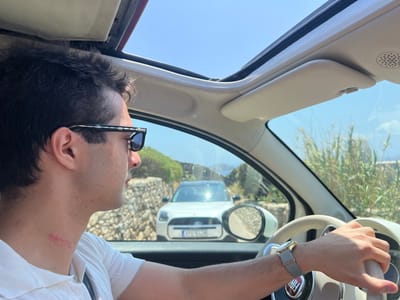 The image depicts the interior of a vehicle with a partially open soft-top roof, positioned on a narrow, unpaved road in Agios Ioannis Diakoftis, Greece, under a clear blue sky.
In the foreground, a male subject with dark, curly hair and wearing dark sunglasses with distinct patterned frames is seated in the driver's position. He is wearing a white collared shirt, and a visible red mark or scratch is present on the right side of his neck. His left hand, adorned with a gold-toned wristwatch with a grey fabric strap, is resting on the light-colored steering wheel, which features a central Fiat emblem. The vehicle's sun visor and a speaker grille are visible on the passenger side above the windshield.
Through the front windshield and driver's side window, the exterior environment is visible. A narrow road, likely dirt or gravel, is flanked by stone walls on both sides. Tall, dry, reedy vegetation grows alongside the road on the right. In the mid-ground, a white crossover SUV, identifiable by its front fascia and what appears to be a Greek license plate (partially visible), is stopped or moving slowly, facing the foreground vehicle. Sunlight strongly reflects off its hood. The landscape beyond consists of low-lying hills or mounds, indicative of a Mediterranean island environment. The overall scene suggests a bright, sunny day.