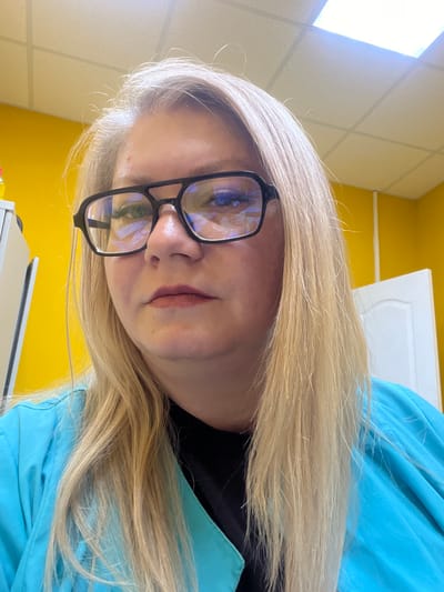 The image depicts a close-up, head-and-shoulders portrait of an adult female individual, positioned indoors in the city of Сундур, Russia. The subject has long, straight, blonde hair and fair skin. She is wearing black-framed eyeglasses with a distinctive double bridge design. Her lips are coated with a red or pink lip product, and her eyes show visible eyelashes. She is attired in a light blue V-neck top, possibly a scrub uniform, over a black undershirt. Her expression is neutral, and her gaze is directed straight at the camera.
The background features bright yellow walls. To the subject's left, a portion of a white cabinet or fixture is visible, along with a partially obscured yellow object. To her right, a white door with a curved panel design is visible against the yellow wall. The ceiling consists of white acoustic tiles, and reflections of overhead fluorescent lighting are present on the subject's eyeglasses and in the upper right quadrant of the image.