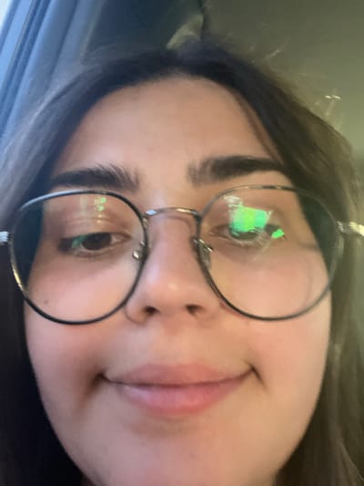The image is a close-up selfie of a young woman with dark hair and glasses. She is wearing round, silver-rimmed glasses with green reflections visible in the lenses. Her eyebrows are dark and well-defined. She has a slight smile on her face, with her lips closed. The background is blurred and appears to be indoors, possibly in a car, with hints of blue and grey tones suggesting daylight. There is no indication of the location being in Luzhany, Ukraine.
