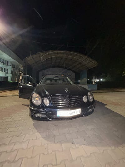 A black Mercedes-Benz E-Class sedan is parked on a paved surface in front of an outdoor stage at night in Dulovo, Bulgaria. The stage has a light-colored roof structure with visible supports. Behind the stage, trees are visible in the darkness. To the left of the stage is a multi-story building with windows. The car's driver-side door is open. The car's license plate reads "CT 0609 PH". The overall lighting suggests nighttime conditions with some artificial light sources present. The ground is made of interlocking paving stones.