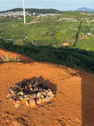 The image depicts a rural landscape in Trường Thọ, Vietnam, characterized by rolling green hills covered in vegetation, likely tea plantations, interspersed with scattered houses and buildings. In the foreground, a circular arrangement of stones sits on a patch of reddish-brown soil, possibly a fire pit. Beyond this, the land slopes upwards, revealing more cultivated hills and a white wind turbine. A town or village is visible on the highest hill in the background, with a dense cluster of houses. The sky is a pale blue, indicating daylight. The scene appears peaceful and natural, with no people or animals present. The overall impression is one of rural tranquility and agricultural productivity.