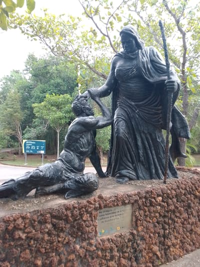 The image shows a bronze statue of two figures in an outdoor setting. One figure, appearing as a woman, stands tall and is draped in a flowing robe, holding a staff. Her hand rests on the head of the second figure, a man who is kneeling and reaching up towards her. The statue is situated on a rough-hewn stone base, with the background featuring trees, foliage, and an overcast sky. A sign in Portuguese is visible to the left of the statue, with the text "PARQUE ESTADUAL MÃE BONIFÁCIA" and other park regulations. This indicates the location is likely the Mãe Bonifácia State Park in Cuiabá, Brazil. The statue depicts a scene of connection or blessing between the two figures.