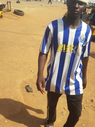 An outdoor, portrait-oriented image captures a dark-skinned individual standing on a dry, sandy, light-brown ground under direct sunlight in Tukur Tukur, Nigeria. The strong, elongated shadows suggest it is either morning or late afternoon.
The subject, likely male, wears a white beanie and a blue and white striped long-sleeved football jersey. On the left chest, a circular crest is visible, identifiable as the emblem of FC Porto, with "FC P" clearly legible within it. Below the collar, several vertical yellow elements, possibly part of a sponsor logo or numbering, are partially obscured and appear worn. The person is also wearing black trousers with white stripes along the side seams. They stand facing slightly towards the camera, with a neutral expression, and their left arm is bent, hand open with the palm facing upwards.
The ground is unpaved and uneven. Scattered objects include a black tire, a metal hoop, pieces of dark plastic or fabric, and a dried plant stem. In the background, an unpaved road stretches into the distance, with vague shapes of vehicles or structures and the legs of another person visible on the far left.