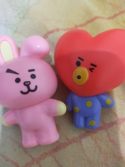 The image shows two small figurines, one pink with rabbit-like ears and the other a red heart-shaped character with a blue outfit adorned with yellow polka dots. The figurines are placed on a patterned surface with pinkish-red markings, possibly a tablecloth or blanket. The lighting suggests an indoor setting. There is no discernible information about the location being in the city of Pematang Siantar, Indonesia.
