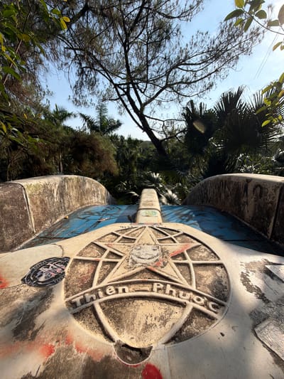 The image shows a close-up of a weathered, likely concrete, structure. The structure features a compass-like design with the words "Thien Phuoc" inscribed in a circle. The design is worn, with some staining and apparent graffiti. The structure is set against a backdrop of lush greenery, including various trees and foliage. The scene is located in Thủy Xuân Ward, Vietnam, as indicated by the inscription on the structure. The view is from a low angle, looking upwards. The sky is partially visible through the trees, appearing bright. There are stickers present on the structure.