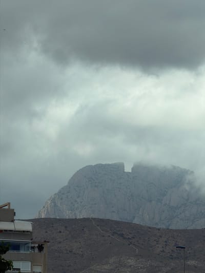 The media file shows a view of a mountainous landscape, possibly in la Vila Joiosa, Spain. The foreground features the top floors of a multi-story building, with modern architectural elements like balconies and large windows. Some vegetation is visible, including what appears to be a palm tree on one of the balconies.
The dominant feature of the image is the imposing mountain range that rises behind the building. The mountains are rugged and rocky, with some sections obscured by mist or clouds. The sky is overcast, with large, grey clouds creating a diffused light. The weather appears to be cloudy or possibly foggy, especially around the mountain peaks.
The time of day is not explicitly indicated, but the soft, diffused light suggests it might be late afternoon or early morning, or simply a heavily overcast day. No people are clearly visible in the image, nor is any specific activity or event taking place. The scene is one of natural grandeur contrasted with human habitation. The muted colors and the presence of mist contribute to a somewhat subdued or atmospheric mood. There is no visible text within the image.