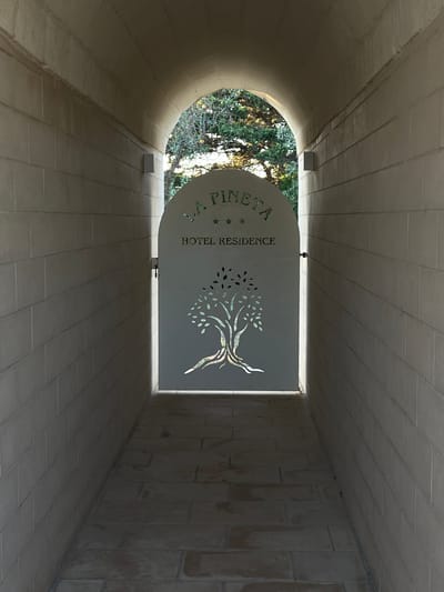 The image shows an arched passageway with light-colored brick walls and a stone-tiled floor. At the end of the passageway, which opens to the outdoors and sunlight, there is a decorative metal gate. The gate is shaped like an arch and features a cut-out design of a tree with spreading branches and leaves. Above the tree design, the words "LA PINETA" are visible, followed by three stars and "HOTEL RESIDENCE". The scene appears to be an entrance or access point to a hotel or residential complex. This location is the entrance to the Hotel La Pineta in Otranto, Italy.