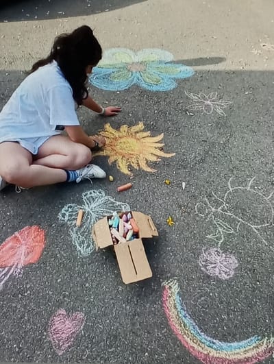 A young adult female is depicted kneeling on a dark asphalt surface, engaged in creating chalk art. She wears a light blue short-sleeved t-shirt, light-colored shorts, and white sneakers with blue socks, and a wristwatch is visible on her left wrist. Her left hand holds a yellow chalk stick, coloring a large, multi-rayed sun design on the ground, while her right hand rests on the pavement for support. Surrounding her are numerous other chalk drawings, including a large light blue and green flower, a red jellyfish, a pink heart, a multicolored rainbow, and several outlined butterflies and smaller flowers. An open cardboard box containing various colored chalk sticks is positioned near her, along with several loose chalk pieces and small yellow petals scattered on the ground. The scene takes place outdoors on a paved area in Ortiugil, Uzbekistan, under natural daylight.