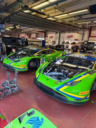 The image depicts a racing garage, likely at the Mugello Circuit in Scarperia, Italy, given the "Mugello Circuit" branding visible on the walls. Two vibrant green Lamborghini race cars, adorned with yellow and blue racing stripes and various sponsor logos, are the primary subjects. The hoods of both cars are open, revealing their engines, suggesting ongoing maintenance or preparation. Several individuals, dressed in casual attire, are present in the garage. Some appear to be mechanics or team members, engaged in activities related to the race cars, such as inspecting or working on them. One individual is looking into the engine bay of the car on the left, while others are gathered around the car on the right. In the background, shelves stocked with equipment, monitors displaying racing data, and garage doors are visible, all contributing to the functional and professional atmosphere of a racing pit stop. The red painted concrete floor of the garage is visible throughout the scene.