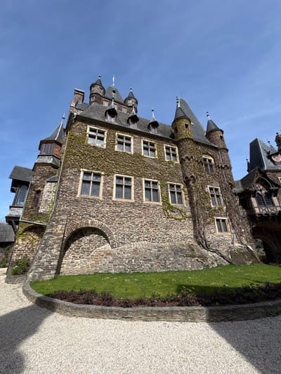 The image shows a building in Cochem, Germany, likely Reichsburg Cochem (Cochem Imperial Castle). The castle is constructed of stone, with multiple towers and turrets visible against a clear blue sky. The walls are partially covered in ivy. Windows are present in rows on the castle walls. At the base of the structure, there is a section of green grass bordered by stonework. Adjacent to the stonework is a gravel path or walkway.