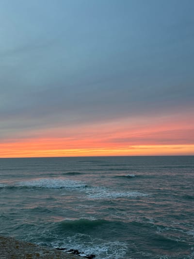 The image presents a panoramic coastal view at either sunrise or sunset, likely from the Atlantic coastline of Cádiz, Spain. The upper half of the frame is dominated by a sky displaying a gradient of colors, transitioning from a pale blue-gray at the top to vibrant horizontal streaks of pink, orange, and yellow hues along the horizon. Below this distinct horizon line, the open ocean occupies the lower half of the image. The water is a deep teal-green color, with numerous waves visible across its surface. White foam crests are prominent on several waves, particularly in the lower left portion of the image, indicating active water movement. A minimal section of the immediate foreground, positioned at the bottom left, shows a weathered, light-colored rocky outcrop or sea wall, with darker rock formations where the water meets the land. No individuals, animals, or man-made objects such as boats are present within the observable scene. The environment is an unpopulated, natural seascape characterized by specific atmospheric lighting.