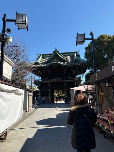 The image depicts a narrow street in Katsushika City, Japan, (latitude: 35.758, longitude: 139.878) with a traditional Japanese building on the left side. The building is a large wooden structure with a green roof and an ornate design. It appears to be a temple or shrine. The street is paved with gray concrete and is lined with buildings on both sides. There is a white tent or awning extending out from a building on the right side of the image. Two streetlights are visible, one on the left and one on the right. A person in a black coat is walking down the street on the right side of the image. There are a few other people visible in the distance. The sky is blue and there are trees visible in the background. The scene is taken from a low angle, looking up at the building. The lighting is bright and the image is clear. The image is likely taken from a mobile phone. There is a black security camera on the streetlight on the right side of the image. The image captures a moment of everyday life in Katsushika City.