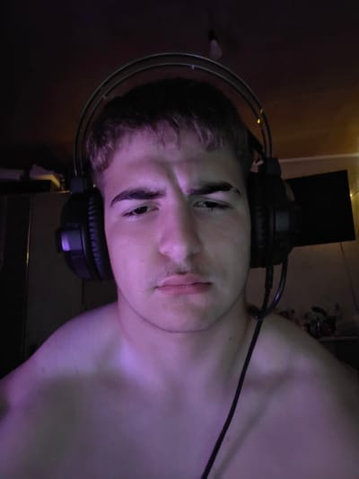 The image shows a close-up, chest-up view of a young male wearing headphones. He is shirtless and illuminated by purple lighting. The background is dark and indistinct, with hints of furniture and a screen on the right. No specific location in Aleksandrovo, Bulgaria is discernible from the image. The man's expression is neutral to slightly concerned. The focus is on his face and upper body.