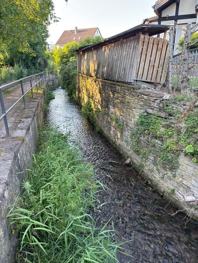 The image depicts a narrow canal or stream flowing through a village. The canal has stone retaining walls on both sides. On the left, a paved path runs alongside the canal, bordered by a metal railing. Lush green grass and reeds grow along the canal's edge. On the right bank, a weathered wooden structure, possibly a shed or covered walkway, is built against a stone wall. The area appears to be a residential or semi-rural part of Bevern, Germany, characterized by greenery, stone architecture, and a small waterway. Sunlight illuminates the scene, casting shadows and highlighting the textures of the stone and wood.