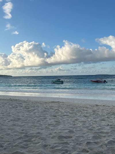 This image depicts a tranquil beach scene with two boats in the ocean. The foreground is dominated by the texture of white sand, showing ripples from the tide. The ocean, a vibrant turquoise and deep blue, stretches towards the horizon, with gentle waves lapping at the shore. Two boats are visible on the water: a smaller green boat is closer to the left side, while a larger red boat with a canopy is situated further to the right. In the distance, a faint landmass can be seen on the left, and the horizon is marked by the meeting of the sea and sky. The sky is a clear blue, dotted with fluffy white clouds, suggesting fair weather. The overall atmosphere is serene and picturesque. The image does not provide enough information to determine a specific location within the city of A R A, Indonesia, or to identify any people or specific actions.