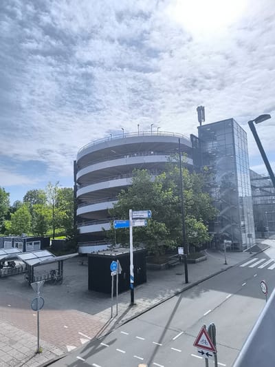 The image depicts a multi-story, circular parking garage located in Hoofddorp, Netherlands, based on the signage. The garage is constructed of concrete and has an adjacent glass-enclosed stairwell with metal stairs visible. A cellular antenna is mounted on top of the stairwell structure. Street signs point towards "Spaarne Gasthuis Hoofdingang" (Hospital Main Entrance) and "Q-Park P+R," and a bicycle route. There's a sheltered bicycle rack area on the left side of the frame. The weather appears overcast, and the surrounding area includes trees and modern office buildings. The street has crosswalks and signage indicating a bicycle crossing with a maximum speed of 30 km/h.