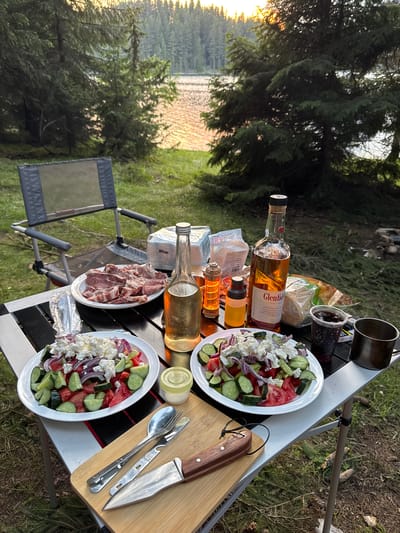 An outdoor picnic scene is set up on a folding table in a wooded area near a lake. The table is laden with food and drinks, including two plates of salad topped with feta cheese, a plate of raw meat, a bottle of Glenfiddich whiskey, and a bottle of clear liquid. There are also small containers, a plastic cup with a dark beverage, a metal mug, and a bag of bread. A folding chair is visible behind the table, and camping gear and firewood are scattered on the ground. The background shows a dense forest and the sun setting over a tranquil lake. The image appears to be taken in the vicinity of Sarnitsa, Bulgaria, given the natural landscape.