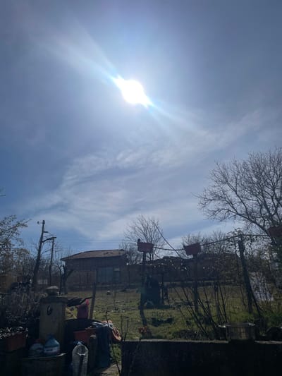 This image captures an outdoor scene in Topchii, Bulgaria, under bright daylight with the sun, displaying a prominent lens flare, positioned in the upper-left quadrant of a partly cloudy blue sky. A faint aircraft contrail is visible in the middle-right sky. The immediate foreground features a dark, low-lying stone or concrete structure. On the left side of this structure, a stone fountain-like element is present, accompanied by multiple plastic water bottles, some identifiable as "DEVIN" brand, a pink bag, discarded fabric, and a red pot. A silver metal pot rests on the structure to the right. The midground consists of a grassy, somewhat overgrown yard or garden area with numerous bare deciduous trees and shrubs, indicating a late winter or early spring season. A two-story house with a red-tiled roof and exposed brick or stone walls is visible in the center-left. Further right in the midground, two tall wooden posts, each topped with a reddish-brown rectangular structure, stand connected by wires or a clothesline. A dark metal drum or container is located near the base of these posts. The background primarily shows additional bare trees and obscured distant structures. No individuals or animals are present.