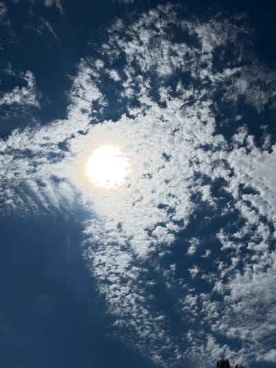 This media file shows a bright, almost blinding sun shining through a scattered cloud cover in a deep blue sky. The clouds are thin and wispy, with some appearing more defined and textured than others, creating a dynamic pattern across the sky. The sun is the dominant element, radiating a strong light that washes out some of the detail in the surrounding clouds, giving the impression of a halo. The overall scene is one of a bright, possibly warm day, with the sun playing hide-and-seek with the clouds. No people, objects, or specific location cues are visible, other than the context of Takhtamukai, Russia. The time of day appears to be mid-day, given the intensity of the sun. The weather is clear but with scattered clouds. No text is visible in the image.