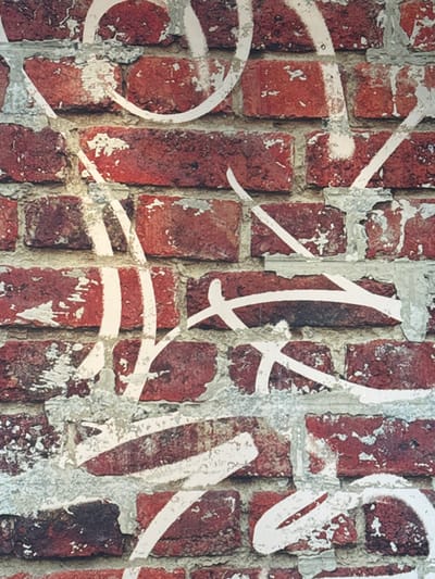A vertical forensic image depicts a section of a weathered brick wall in Oberhausbergen, France. The wall is constructed from rectangular red-brown bricks, exhibiting variations in color, surface wear, and minor chipping. The mortar between the bricks is a light gray, appearing unevenly applied, eroded, and peeling in numerous areas, revealing rough textures and some underlying brick edges. Superimposed across the entire wall surface is extensive graffiti executed in white spray paint. This graffiti consists of thick, fluid, abstract lines, curves, and strokes, including a partial circular element in the upper left, various diagonal lines, and horizontal wavy forms. The white paint application is somewhat inconsistent in opacity, suggesting multiple layers or varying spray pressure. There are no individuals, other distinct subjects, or specific actions identifiable within the frame beyond the static architectural element and its markings. The environment is limited to this detailed wall section.