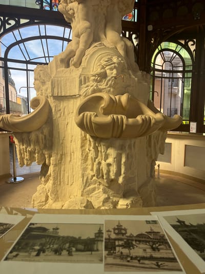 The image shows a close-up of a large, light-colored stone fountain sculpture inside a building. The sculpture depicts several figures, including what appears to be a cherubic form at the top, and carved faces and shell-like basins lower down. The stone has a rough, textured finish, resembling natural rock formations with some drips or icicle-like elements. The sculpture is lit from the front, casting shadows that highlight its details.
Behind the sculpture, large arched windows with dark metal frames overlook an exterior scene. The windows have stained glass elements in the upper portions, with colorful geometric patterns and floral motifs. Through the windows, buildings with balconies and what appears to be a street scene can be glimpsed, with hints of blue sky and clouds. The interior setting includes wooden architectural elements and a low wall or counter in the background.
In the foreground, several black and white historical photographs are laid out on a surface. These photographs display street scenes with ornate buildings, people, and possibly fountains or monuments. The photographs are slightly blurred due to their distance and the focus of the main image.
The context provided by the question indicates this scene is located in the city of Évian-les-Bains, France. Given the architecture, the fountain's style, and the historical photographs, it is likely this is an interior view of a public building or establishment in Évian-les-Bains, possibly a spa, casino, or municipal building, showcasing a significant piece of decorative art. The overall impression is one of historical grandeur and artistic display.