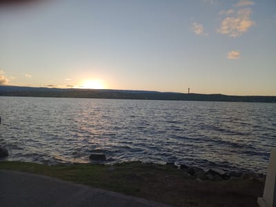 The image presents a serene sunset over a large body of water, consistent with Lake San Roque in Villa Carlos Paz, Argentina. The sun, a brilliant orb, is positioned low on the horizon, partially hidden by the distant hills and casting a strong, warm glow across the scene. Its light creates a shimmering path of reflection on the gently rippling water surface.
The sky transitions from a soft yellow-orange near the horizon to a clear blue-grey higher up, dotted with a few wispy clouds that catch the last rays of sunlight. Across the water, a distant shoreline reveals rolling hills or mountains, upon which a town or city appears to be built. A notable detail on the right side of the distant hilly horizon is a tall, dark, cylindrical structure, possibly a tower or monument, standing out against the twilight sky.
In the foreground, a strip of land borders the water. This area consists of dark earth and sparse grass, with several rocks along the water's edge. A paved path or sidewalk runs along the bottom left of the frame, suggesting a promenade or viewing area. No people or specific activities are visible, emphasizing the tranquility of the natural landscape during dusk. There is no visible text in the image.
