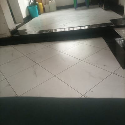 The scene depicts an interior view within the city of Kimorori/Wempa ward, Kenya. The floor in the foreground and midground is covered with light-colored, possibly off-white or light grey, square ceramic tiles laid in a diagonal pattern with dark grout lines. A pronounced, raised section of black tiles, approximately two tile heights high, traverses the midground horizontally, segmenting the floor. This black-tiled section then turns and extends perpendicularly towards the right, forming a barrier or change in floor level. Beyond the raised black tiles, the floor continues with the same light-colored diagonal tiling. Along the back wall, from left to right, a light blue plastic bucket is visible, followed by a yellow plastic container, and then three translucent or white rectangular plastic jerrycans. In the far right background, two objects exhibiting green and blue coloration, possibly parts of chairs or utility items, are partially visible, alongside a darker, indistinct object. No individuals, actions, or interactions are observed within the image.