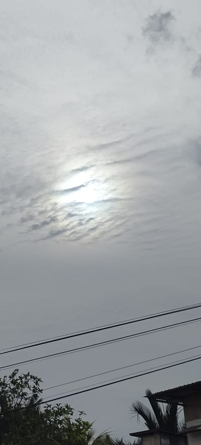 The image shows a cloudy sky with the sun partially visible through the clouds, creating a bright halo effect. In the foreground, there are several power lines stretching horizontally across the frame. Below the power lines, the tops of trees and the roof of a building are visible. The scene appears to be an outdoor view, possibly from a residential area. The location is identified as Meunasahceubrek, Indonesia.