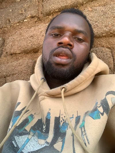 A medium shot photograph shows a Black man in a tan hoodie against a textured, light brown brick wall. The man is positioned centrally in the frame, facing the camera directly with a neutral expression. His head is slightly tilted, and his eyes are open, looking forward. He has short, dark hair and a short beard and mustache. The hoodie is a light tan color with a graphic print on the front featuring stylized black and blue imagery. A light tan drawstring with a metal eyelet is visible on the left side of the hoodie. The brick wall behind him is rough and uneven, with visible individual stones and mortar. The lighting suggests an outdoor environment. The specific location of Buga da Gindi, Nigeria is not ascertainable from the image itself.
