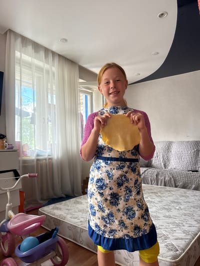 A young girl with blonde hair and fair skin, wearing a floral apron and a pink knitted sweater, stands in a room and holds a round, flat piece of dough. She is smiling directly at the camera. Behind her, sheer white curtains cover a large window. To the left, a white dresser and a child's tricycle are visible. In the background, a gray couch and a mattress lie on the floor. The room has a high ceiling with recessed lighting. The setting is identified as a residential interior in the city of Valday, Russia.