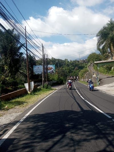 The image depicts an asphalt road ascending through a lush, hilly, and vegetated landscape in the city of Ban Phonhxavanh, Laos, under a blue sky with scattered white cumulus clouds. Dappled shadows from trees and utility poles are cast across the road surface. Multiple dark-colored utility poles, densely strung with numerous black wires, line the left side of the road, intertwined with abundant green foliage and tall trees. On the left, a large rectangular billboard displays advertisements for "EMBAH TERIAI RESORT" and "PANAS," with visible phone numbers (0811 205 219, 0263 512 505) and the word "Cianjur" alongside a logo. A white, reflector-like object is positioned on the grassy verge beneath the billboard. The road features a solid white line on its right edge and a broken white center line. Several individuals on motorcycles and scooters are visible, traveling uphill away from the viewer. The lead vehicle is a blue scooter with a single rider wearing a white helmet. Behind it, a lighter-colored scooter carries at least one rider, possibly two, with a red and white helmet visible. Further along the winding road, a line of additional motorcycles proceeds uphill. On the right side of the road, a smaller, green-roofed structure or sign is partially visible amidst the dense vegetation, and another road branches off uphill to the right. Hillsides covered in dense trees and scattered buildings are present in the distant background.