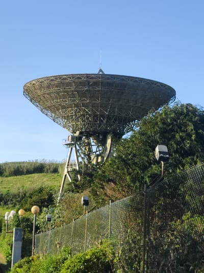 A large, greyish-metallic parabolic dish antenna, characteristic of a radio telescope or satellite dish, dominates the central and upper-left portion of the image. The dish features a visible metal framework on its underside and is supported by a substantial metal truss structure with multiple support legs. A central mast, likely housing a feedhorn or primary antenna element, extends from the dish's focal point.
The structure is situated on a green, undulating hillside covered with dense vegetation, including various shrubs, trees, and tall grasses. The sky above is clear and light blue. In the foreground, a chain-link fence, topped with what appears to be a deterrent wire, runs horizontally from the left to the right side of the frame, partially obscured by green foliage. Several outdoor lighting fixtures are positioned along the fence line: some are spherical and pale yellow/white, while others are rectangular and dark grey/black, mounted on curved poles above the fence. This scene is located in Ponta Delgada, Portugal. No human figures or animals are visible.