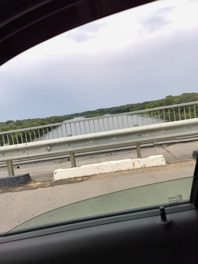 The image shows a view from a car window, looking out over a bridge. The bridge has a metal railing and a paved road surface. Beyond the bridge, a wide river flows, with lush green trees lining its banks. The sky is overcast with gray clouds. The foreground shows part of the car's interior, including the window frame, a portion of the door panel, and a black door lock. There are no people visible in the image. The scene appears to be a serene landscape, possibly on the outskirts of Belogorskiy, Russia, given the context.